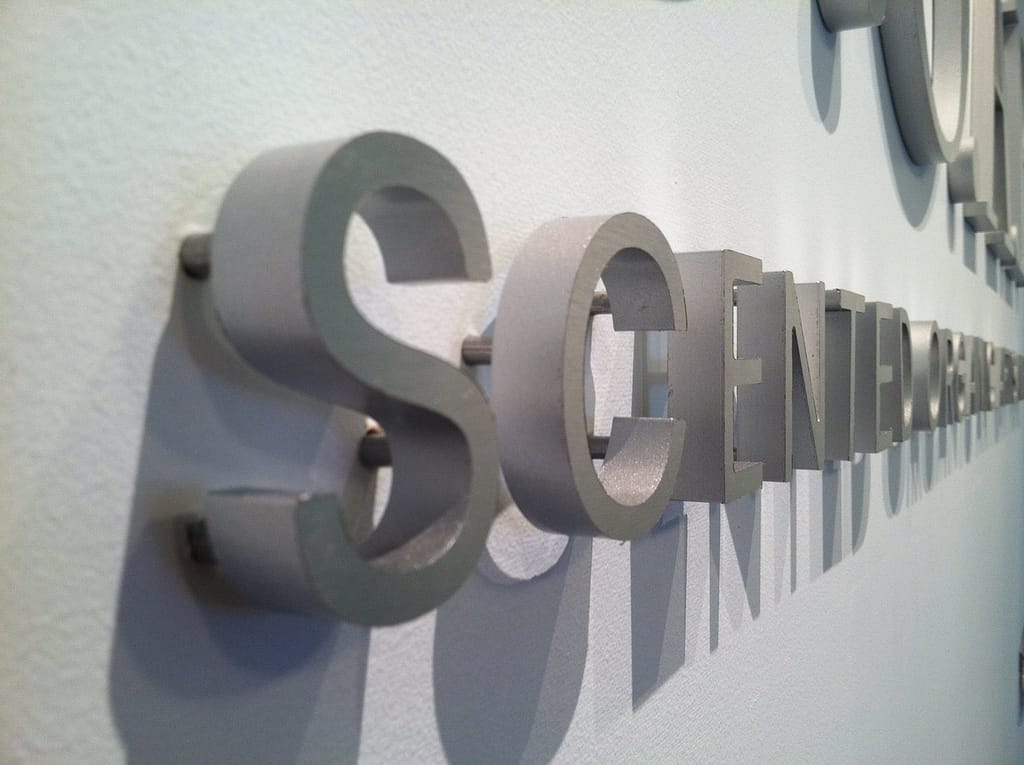 Close up of a aluminum sign mounted to white painted drywall. The sign is 1/2" thick and is pin mounted with 1/4" standoffs.