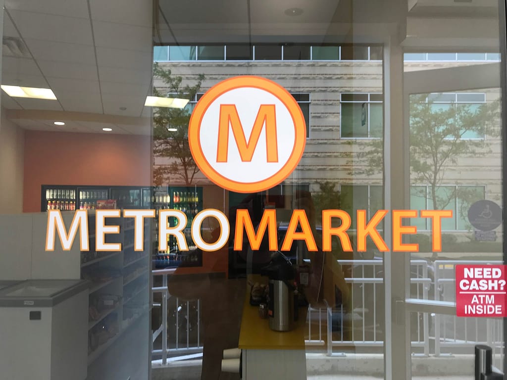 Full color glass graphic. An orange and white circle with a M in the center. Below it is the text in orange and white saying "METRO MARKET"