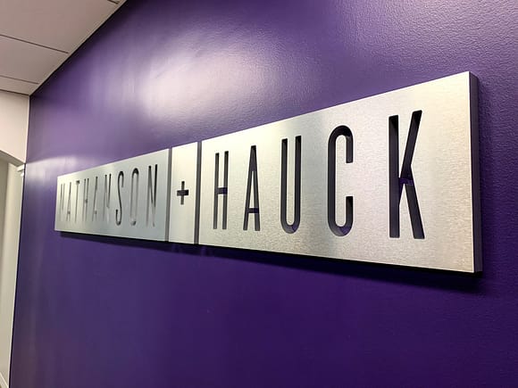 Laminate on acrylic sign mounted to a purple wall. The sign is 1/2" thick with a brushed aluminum laminate applied to the face. The letters "Nathanson + Hauck" is cut out.