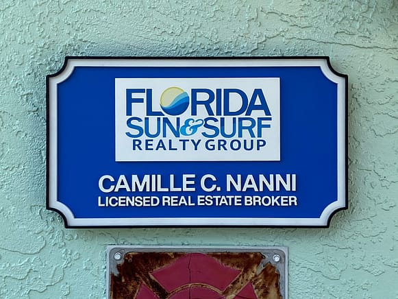 Blue, white and black wood realty sign. The sign is multilayered and multicolored. The center has "Florida Sun & Surf Realty Group" and below is the brokers name