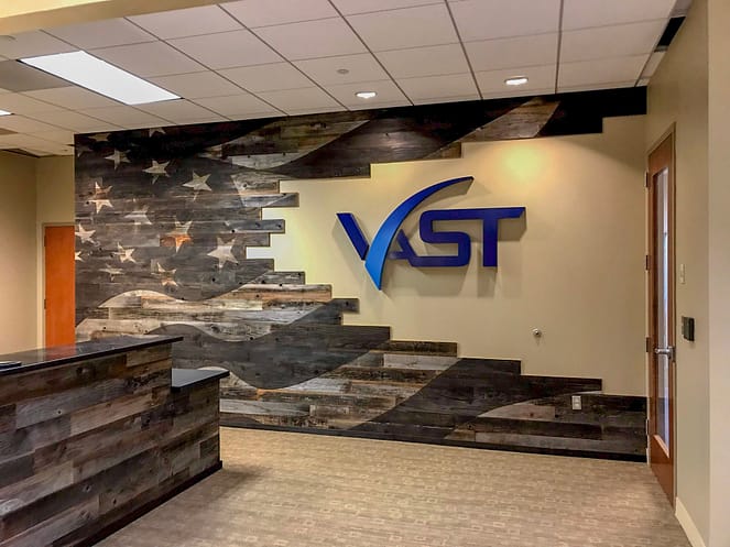 A reception area with an American flag stained into the reclaimed wood on the wall. The desk also has been wrapped in reclaimed wood. There is also a sign on the wall saying "Vast".